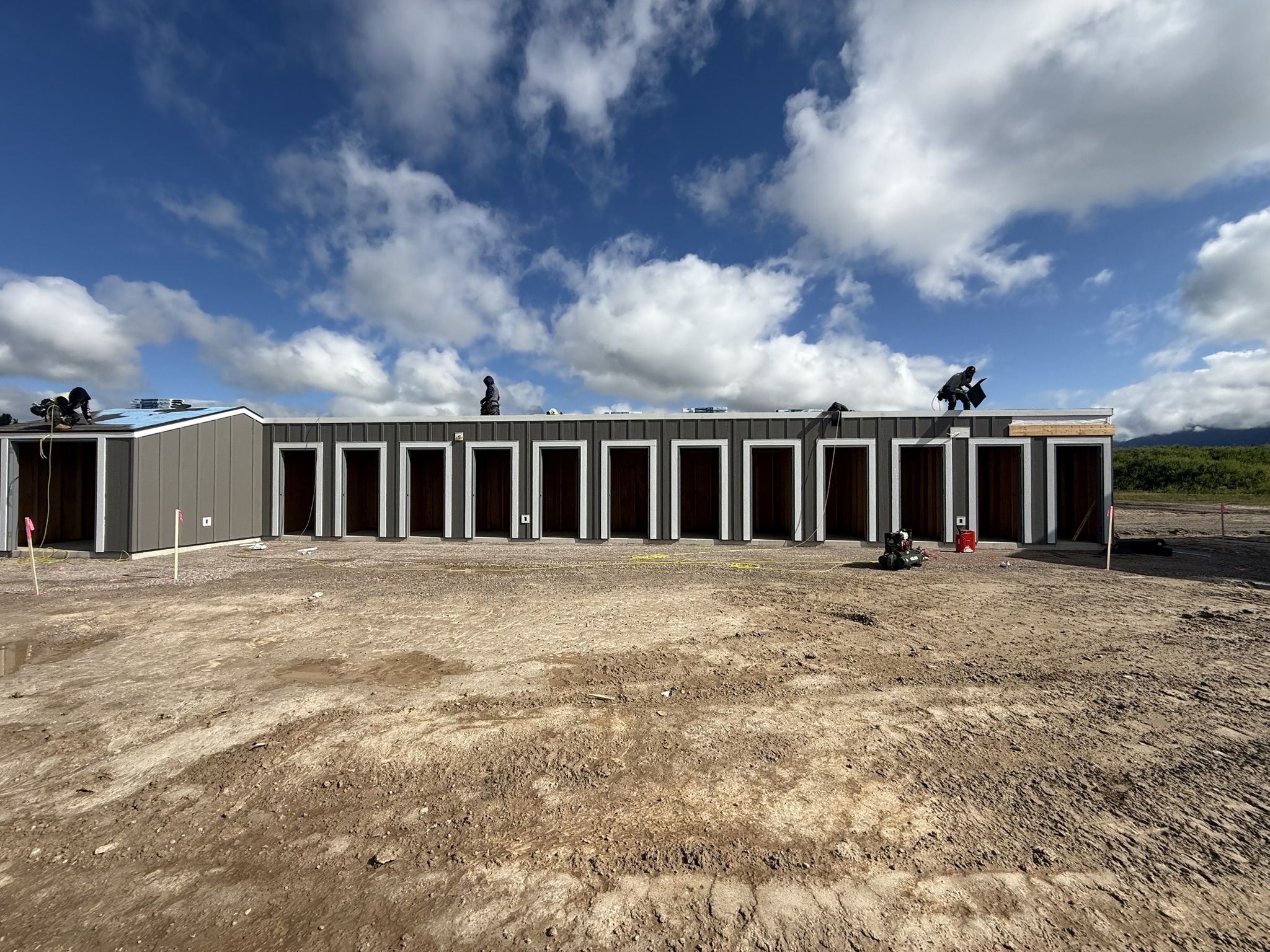 Long metal storage-unit building with crews working the roof on a graded dirt lot