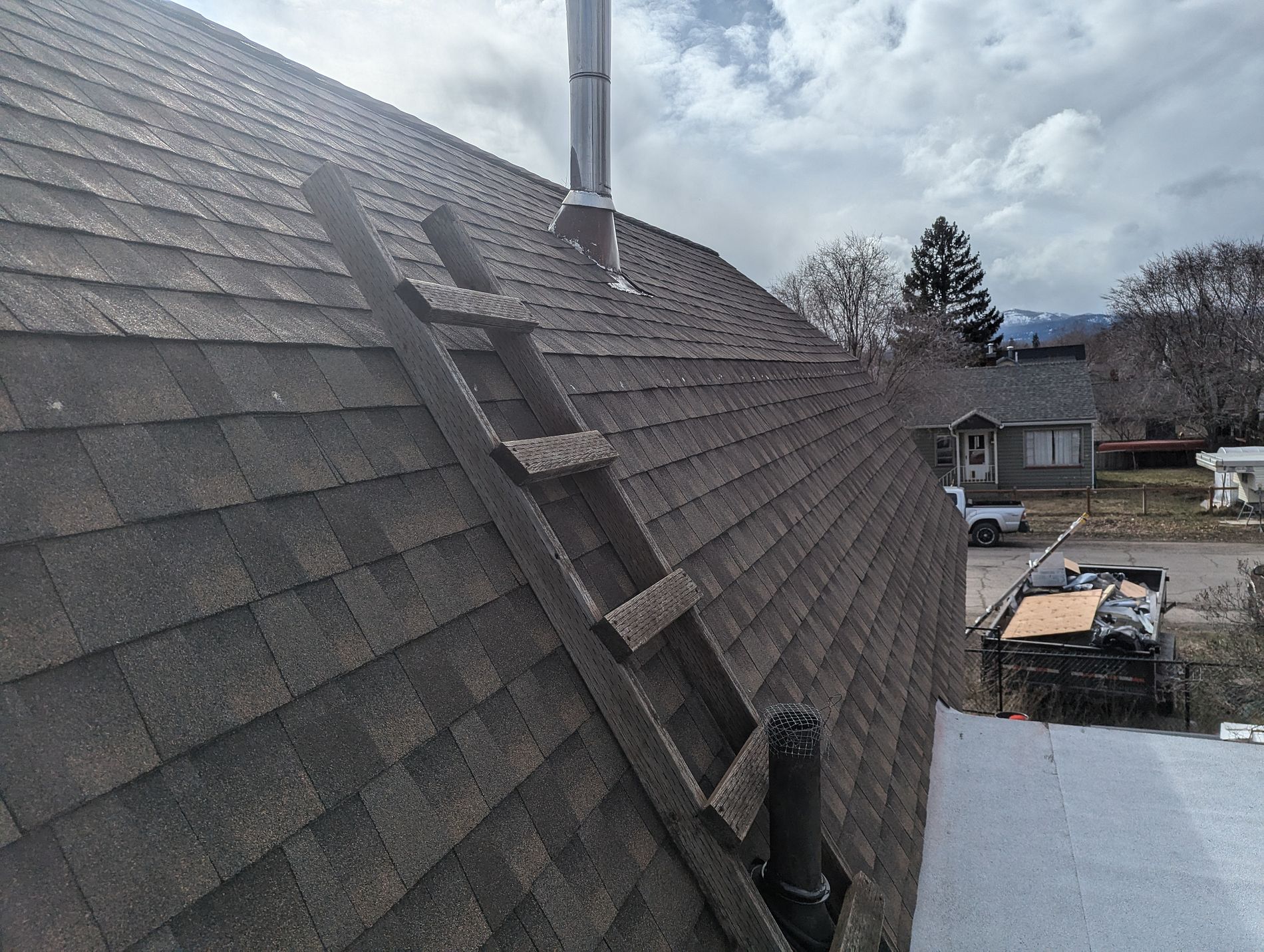 Architectural asphalt shingle roof with a metal furnace flue and a roof ladder, homes and hills beyond.