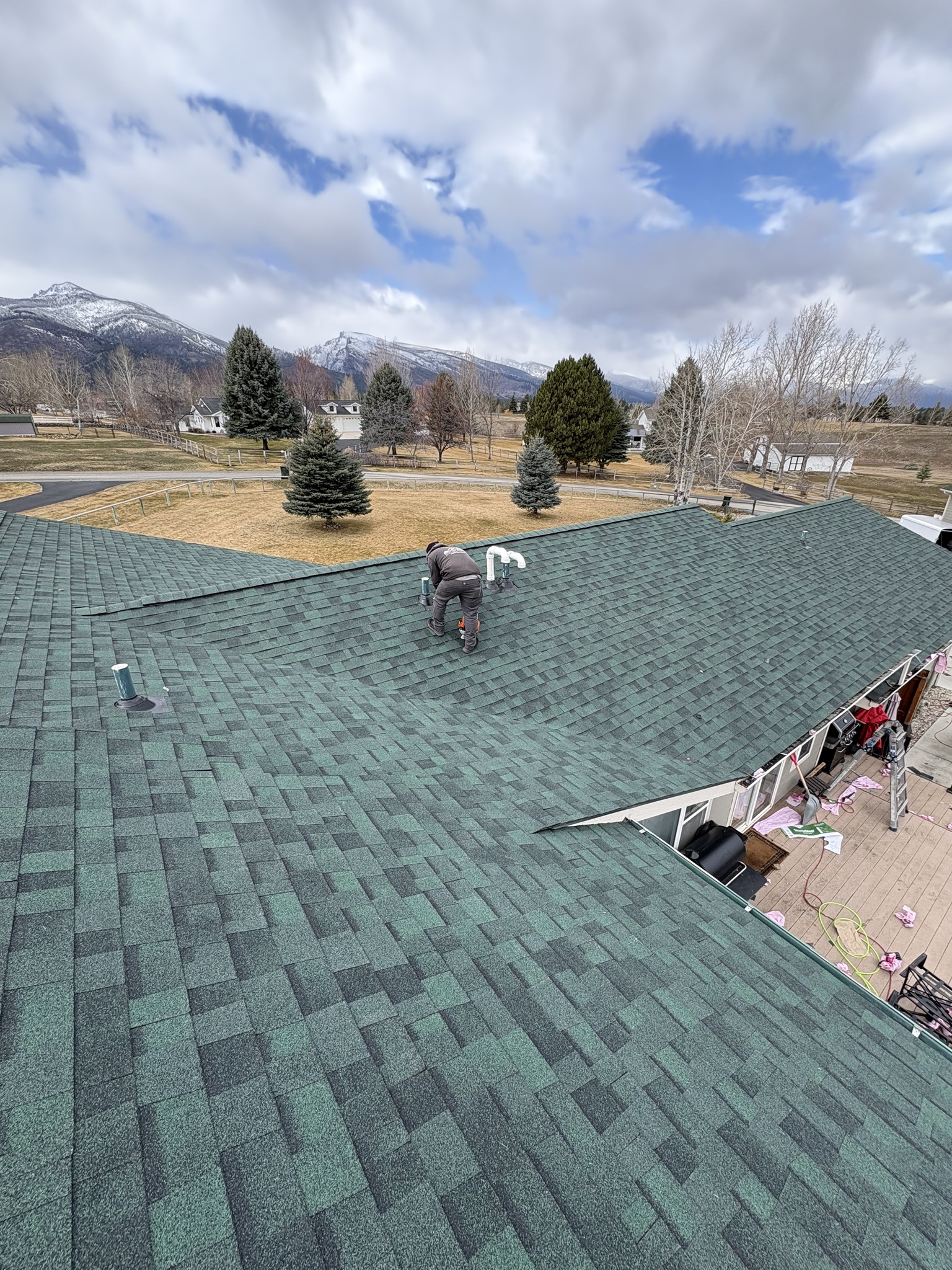 Green architectural shingle roof under installation with a roofer at work and mountains behind